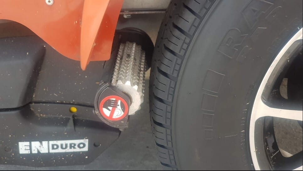 카라반 타이어 측면에 위치한 엔듀로(ENDURO) 무버 장치. 거친 톱니바퀴 모양의 금속 구동 롤러가 타이어 표면에 닿기 직전의 근접 사진이며 손 끼임 주의 경고 마크가 보임.