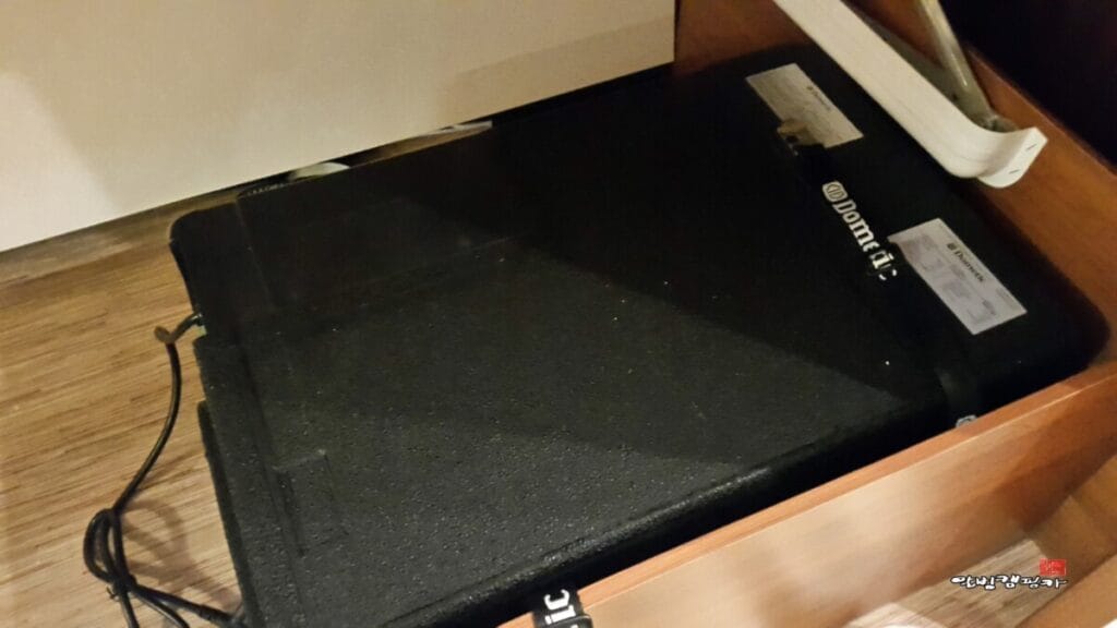 타공된 수납장 공간에 딱 맞게 안착된 검은색 도메틱(Dometic) 언더벤치 에어컨 본체 모습. 제품 스티커가 붙어있고 전원 케이블이 밖으로 빠져나와 있습니다.